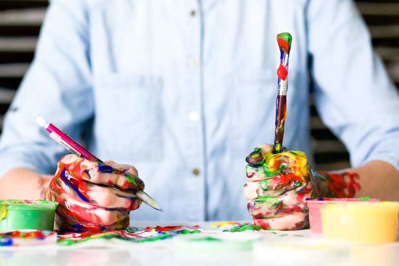 A person in a button up shirt has messy hands that are holding paint brushes